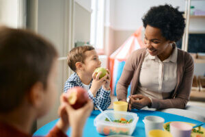 Happy boy eating apple while communicating with his teacher during lunch time.